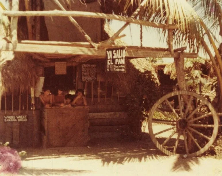 An old film photo of a palm-thatched bakery stand in San Blas, with kids leaning on the wooden counter under a “Ya salió el pan” sign and “Whole Wheat Banana Bread” lettered on the front