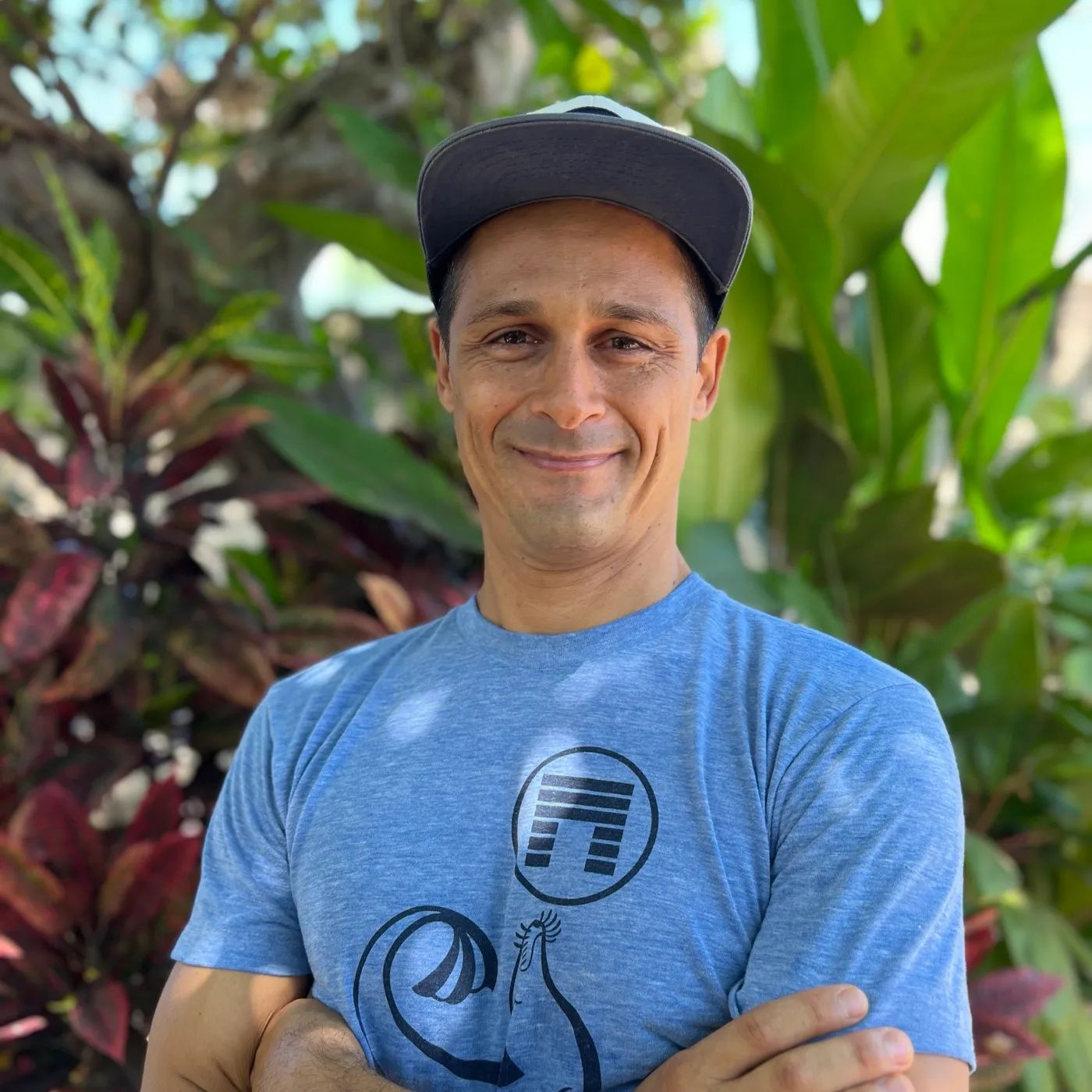 Gibran Garcia outdoors among tropical plants, smiling, arms crossed, in a cap and a blue t-shirt