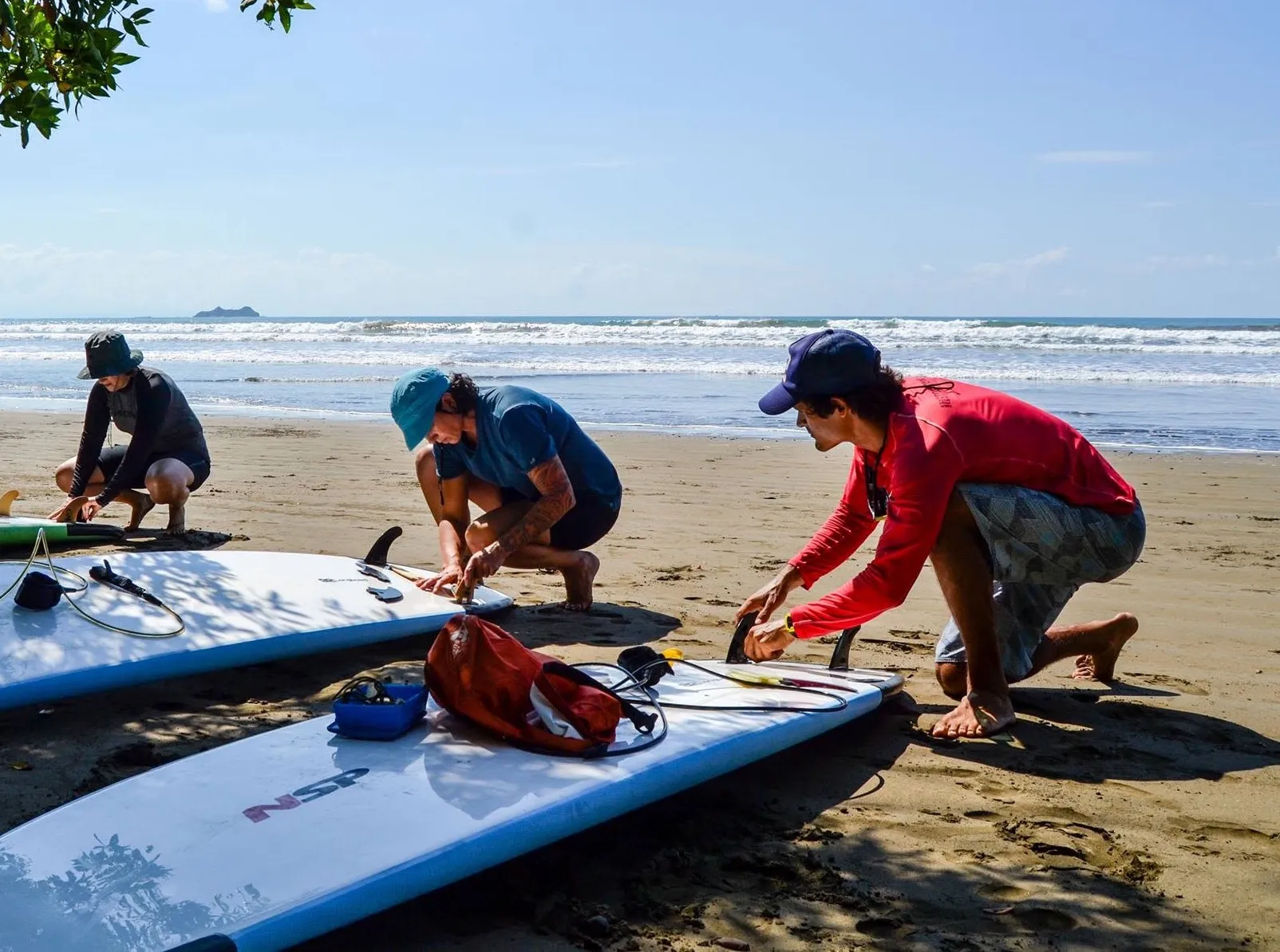 Three surf instructors kneeling on a Costa Rica beach, setting up surfboards at the water's edge